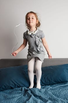 Little girl singing holding headphones cord imitating herself a real singer. Child having fun jumping dancing listening to music on bed in bedroom at home