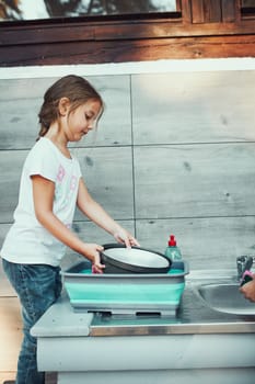 Teenager girl washing up the dishes pots and plates with help her younger sister in the outdoor kitchen during vacations on camping. Camp life. Sisters spending time and working together