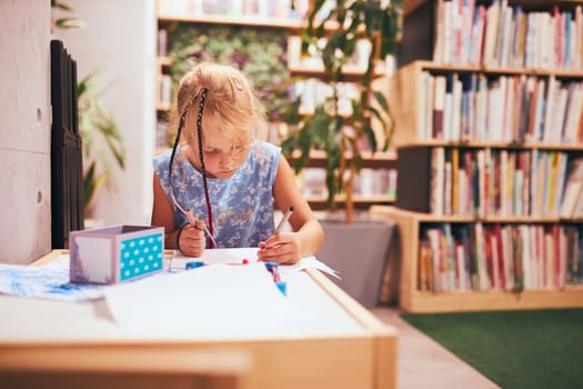 Schoolgirl doing homework. Elementary student learning, drawing pictures, doing puzzles at desk in school library. Primary student in school club. Smart girl learning from book
