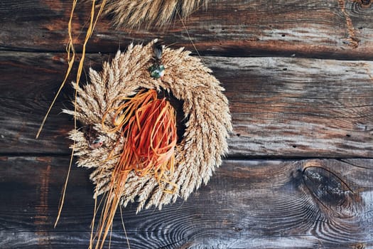 Wreath of golden ears of wheat, dried flowers and herbs tied with red ribbon hung on wooden wall. Natural rustic decoration