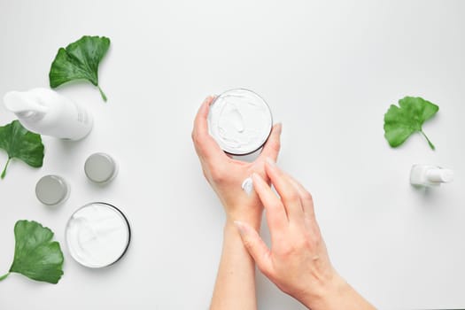 Woman applying  cosmetic moisturizing hand cream. Cosmetic products, green leaves on white table. Spa, manicure, skin care concept. Flat lay, overhead view