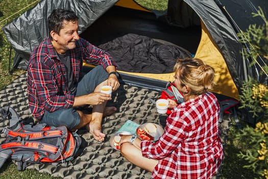 Couple having breakfast sitting by tent at camping. People actively spending summer vacations close to nature outdoors. Concept of camp life