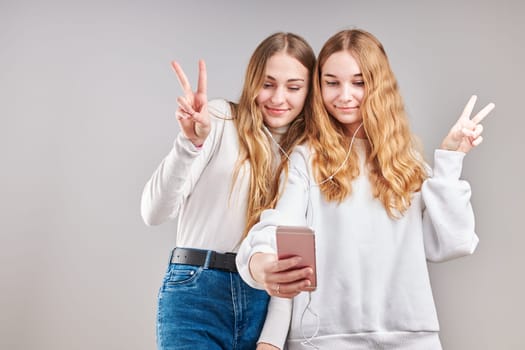 Young women girls listening to music together streaming content having fun watching video enjoying video chat talking with friends making gestures faces using smartphone earphones headphones standing over plain grey background