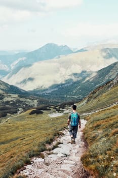 Young man with backpack hiking in a mountains, actively spending summer vacation. Rear view of teenager walking down from top of a hill to valley along mountain footpath