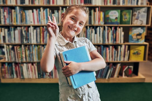 Back to school. Happy little girl at school again. Smiling student making V sign hand gesture holding books standing at front of bookshelf with books in classroom at primary school. Child having class. Positive attitude. Schoolgirl learning at primary school. Elementary education