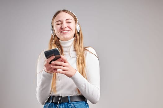 Young woman girl listening to music streaming content having fun watching video enjoying video chat talking with friends making gestures faces using smartphone earphones headphones standing over plain grey background