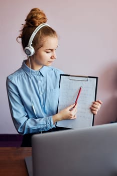 Young woman student showing homework, having classes, learning online, watching lesson remotely, listening to professor, talking with classmates on video call from home during quarantine. Young girl using laptop, headphones, books, manuals sitting at a desk