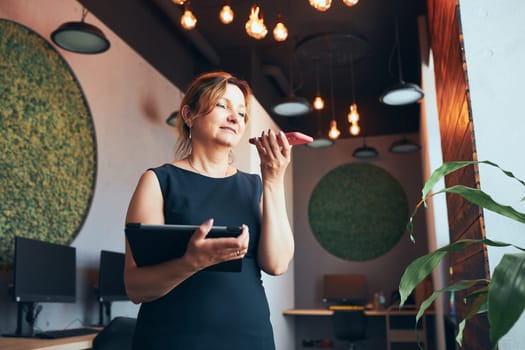 Businesswoman having business call working on tablet in office. Mature busy woman using touch pad computer standing by window in modern interior. Female manager focused on work holding digital device. Using technology