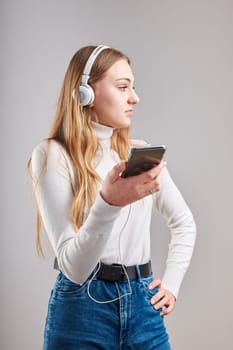Young woman girl listening to music streaming content having fun watching video enjoying video chat talking with friends making gestures faces using smartphone earphones headphones standing over plain grey background