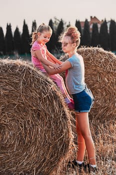 Sisters, teenage girl and her younger sister playing together on hay bale outdoors in the field in the countryside. Candid people, real moments, authentic situations