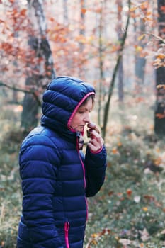 Woman smelling a mushroom picked in forest. Forest in autumn season. Colorful foliage on trees lit by morning sunlight. Natural nature forest landscape in autumn warm sunlight day. Real people, authentic situations