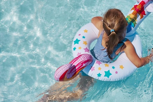 Little girl playing in a swimming pool with inflatable rubber ring toy in the shape of unicorn. View from above of kid in a turquoise water swimming in rubber colorful ring