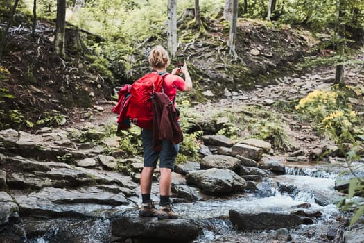 Taking pictures from vacation. Woman with backpack taking photos of landscape using smartphone camera standing on rock on mountain stream during summer trip
