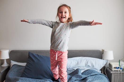 Little happy playful kid girl having fun making silly funny faces jumping on bed in bedroom in the morning at home