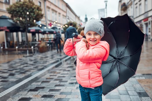 Little smiling happy girl holding big umbrella walking in a downtown on rainy gloomy autumn day