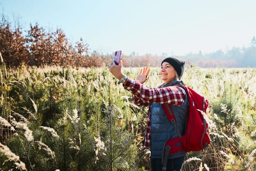 Woman waving during video call sending greetings from vacation trip in mountains. Woman with backpack hiking through tall grass along path on meadow. Spending summer vacation close to nature