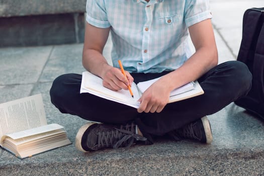 Student making the notes learning from books sitting on a monument outside of university. Young boy wearing a blue shirt and dark jeans