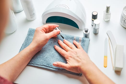 Woman applying gel hybrid polish using UV lamp. Beauty wellness spa treatment concept. Cosmetic products, UV lamp, green leaves on white table. Spa, manicure, skin care concept. Flat lay, overhead view