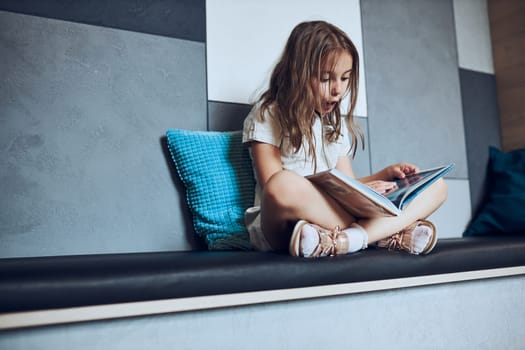 Surprised schoolgirl looking at book page. Child reading book in school library. Primary school pupil is involved in book. Child doing homework. Smart girl learning from book. Benefits of everyday reading. Child curiosity