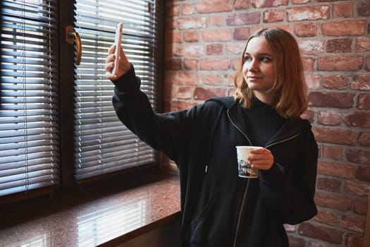 Young woman having video call, talking remotely, taking selfie photo holding smartphone. Girl drinking coffee relaxing while taking break in office standing at window