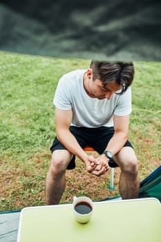 Young man drinking a coffee sitting at front of tent in the morning. Teenager enjoying free time during weekend trip in summer