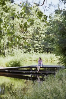 Woman sitting on a bridge over a lake, among the trees, close to nature, during summer vacations. Candid people, real moments, authentic situations