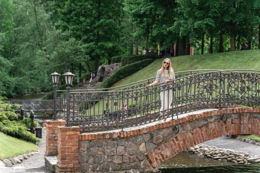 A woman stands on an old stone bridge with a forged fence in the park in the summer.