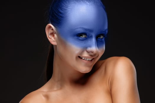 Portrait of a smiling young woman who is posing covered with blue paint in the studio on a black background.