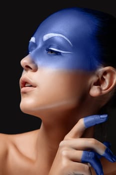 Portrait of a young woman who is posing covered with blue paint in the studio on a black background. girl&#39 s hand around the face and painted blue