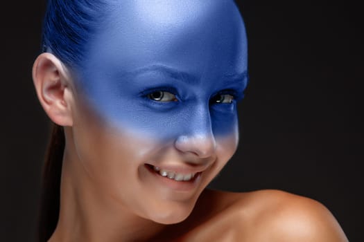 Portrait of a smiling young woman who is posing covered with blue paint in the studio on a black background.