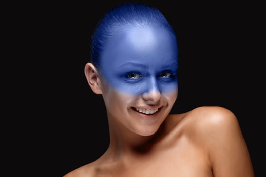 Portrait of a smiling young woman who is posing covered with blue paint in the studio on a black background.