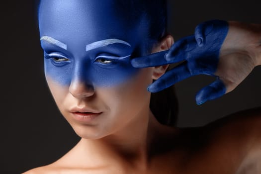 Portrait of a young woman who is posing covered with blue paint in the studio on a black background. girl&#39 s hand around the face and painted blue