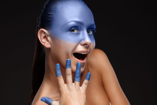 Portrait of a smiling young woman who is posing covered with blue paint in the studio on a black background. girl&#39 s hand around the face and painted blue