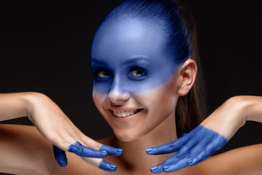 Portrait of a smiling young woman who is posing covered with blue paint in the studio on a black background. girl&#39 s hands around the face and painted blue