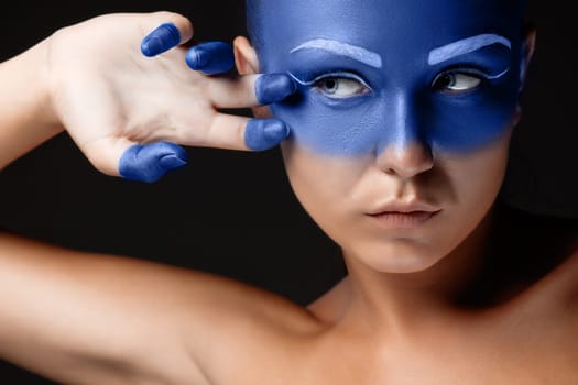 Portrait of a young woman who is posing covered with blue paint in the studio on a black background. girl&#39 s hand around the face and painted blue