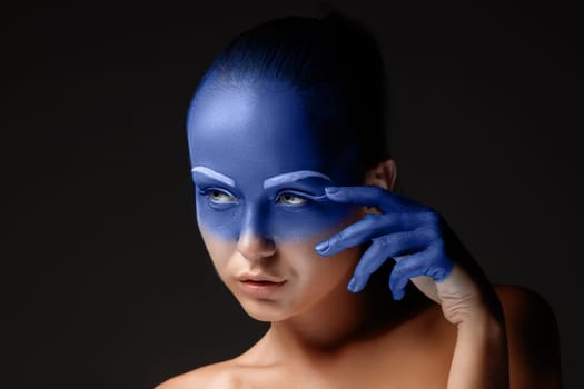 Portrait of a young woman who is posing covered with blue paint in the studio on a black background. girl&#39 s hand around the face and painted blue