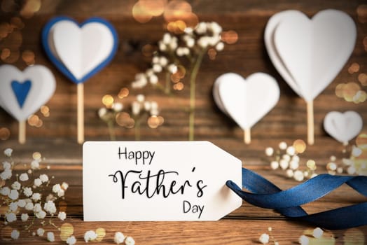 Label With English Text Happy Fathers Day. White Festive And Atmospheric Decoration Like Hearts, Flowers And A Blue Bow. Vintage, Wooden Background With Sparkling Light.