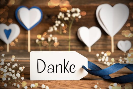 Label With German Text Danke Means Thank You. White Festive And Atmospheric Decoration Like Hearts, Flowers And A Blue Bow. Vintage, Wooden Background With Sparkling Light.