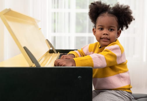Curly girl playing piano and looking at camera. Side view kid sitting with piano. Music education and leisure activity concept.
