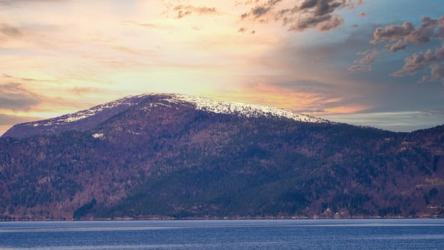 Fjord with snow covered mountains on horizon and sunset with shining clouds. The water glitters in Norway. Landscape photo from the north