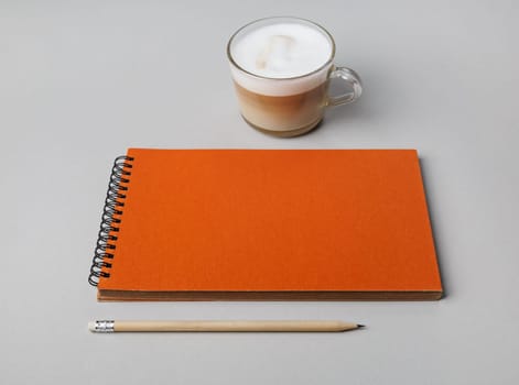 Blank orange notebook, pencil and coffee cup on gray paper background. Responsive design mockup.