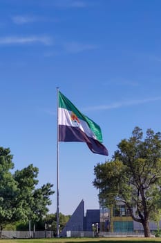 Merida, Spain - April 17th, 2023: Extremadura Region flag waving over blue sky. Lusitania Bridge roundabout, Merida, Spain