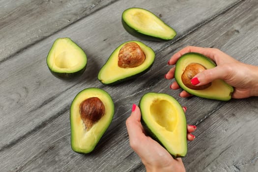Avocado halves on gray desk, woman hands with red nails hold two of them.