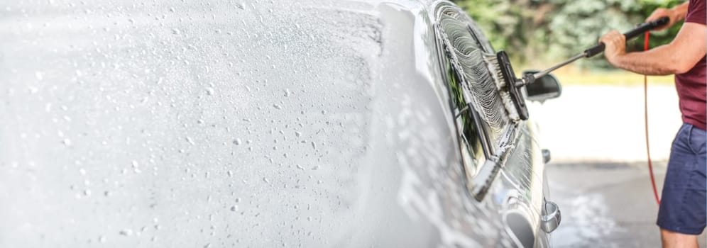 Man washing his car at self serve carwash, brush cleaning glass covered in shampoo. Left side is expanded to banner size, space for text.