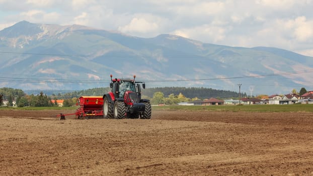 Tractor sowing on empty field, village houses and mountains in background.