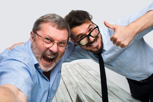 Two smiling colleagues taking the picture to them self sitting in the office, happy friends with glasses taking selfie with telephone camera  on white background