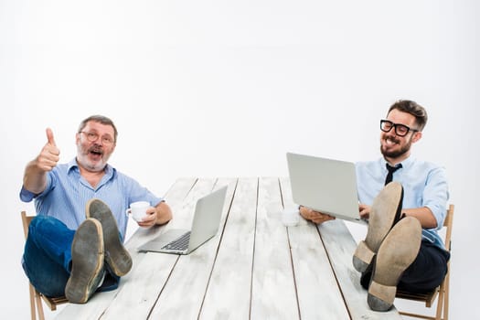 The two smiling businessmen with legs over table working on laptops on white background. Business in the American style