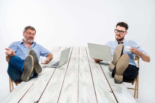 The two smiling businessmen with legs over table working on laptops on white background. Business in the American style