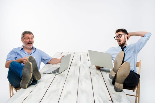 The two smiling businessmen with legs over table working on laptops on white background. Business in the American style