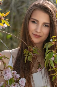Beautiful long haired brunette girl is posing on camera outdoors covered by sprig of sakura tree. Vertically.